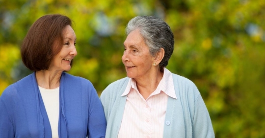 elderly mother with her daughter standing outdoors smiling at each other.